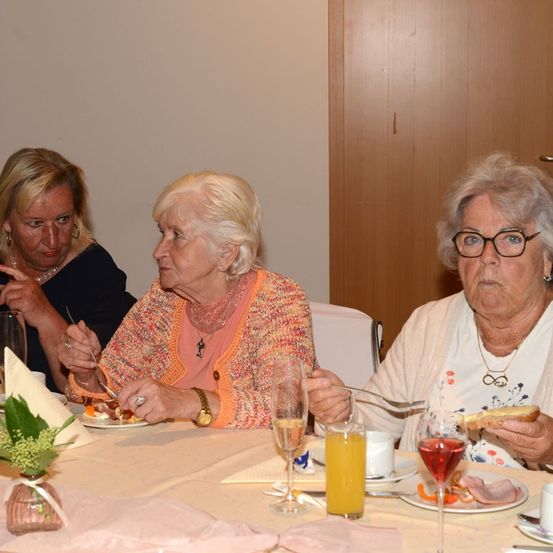 Drei ältere Frauen sitzen an einem Tisch, jede mit einem Teller Essen und einem Glas Wein. Eine Frau trägt eine Brille und eine Halskette, eine andere eine Uhr.