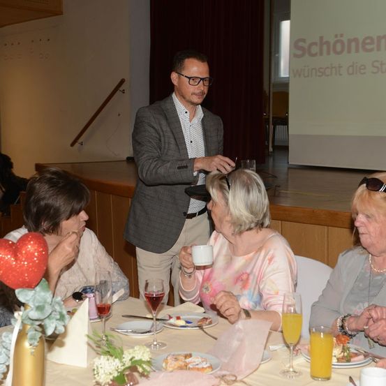 Ein Mann in einem Anzug steht an einem Tisch und spricht zu einer Gruppe von Frauen, die sitzen und essen. Eine Projektionsleinwand hinter ihm zeigt eine Nachricht auf Deutsch.