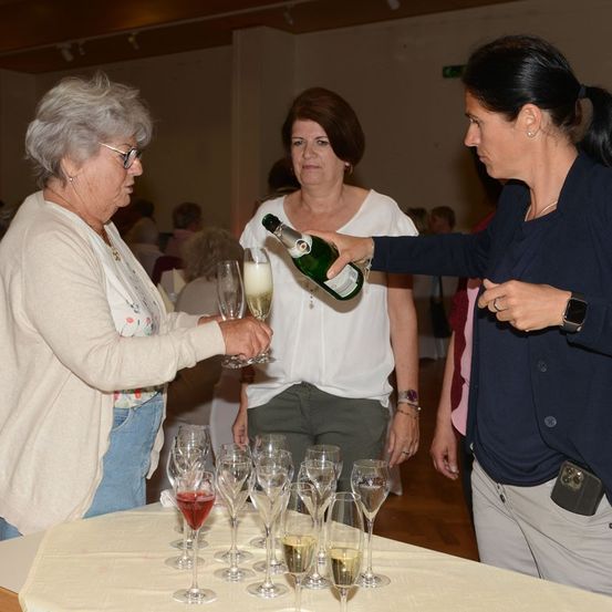 Drei Frauen stehen um einen Tisch mit Weingläsern. Eine Frau schenkt Wein aus einer Flasche in ein Glas, das eine andere Frau hält.