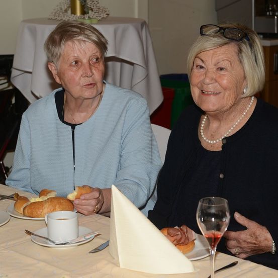 Zwei ältere Frauen sitzen an einem Tisch, eine mit Croissant und Kaffee, die andere mit einem Schinkensandwich und Wein. Beide lächeln.