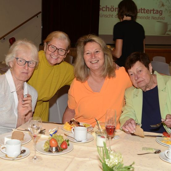 Vier Frauen sitzen an einem Tisch, lächeln und posieren für ein Foto. Sie sind umgeben von Tellern mit Essen, Weingläsern und einer Blumenvase. Im Hintergrund sind eine Treppe und eine Projektionsleinwand zu sehen.