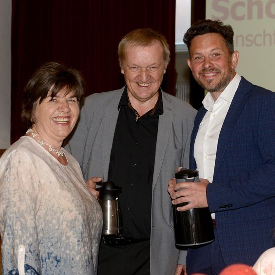 Drei Personen stehen zusammen, lächeln und halten Kaffeebecher. Der Mann links trägt einen grauen Blazer, die Frau links hat eine Halskette und der Mann rechts ist in einem blauen Anzug.