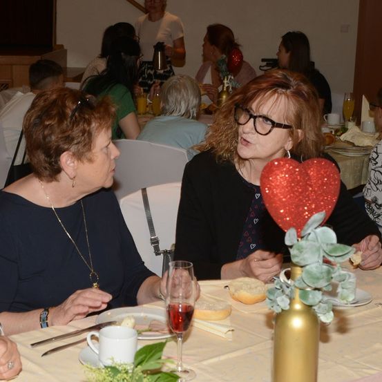 Zwei Frauen sitzen an einem Tisch, eine mit einer Brille und einer Halskette, die andere mit einem herzförmigen Luftballon. Sie sind von anderen Menschen umgeben, die an einem Tisch essen.