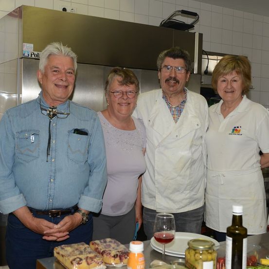 Vier Personen stehen lächelnd in einer Küche für ein Foto. Ein Mann in einem Jeanshemd und eine Frau in einem weißen Hemd sind links, während ein Mann in einem weißen Kochmantel und eine Frau in einer weißen Schürze rechts sind. Sie sind von verschiedenen Küchenutensilien umgeben.