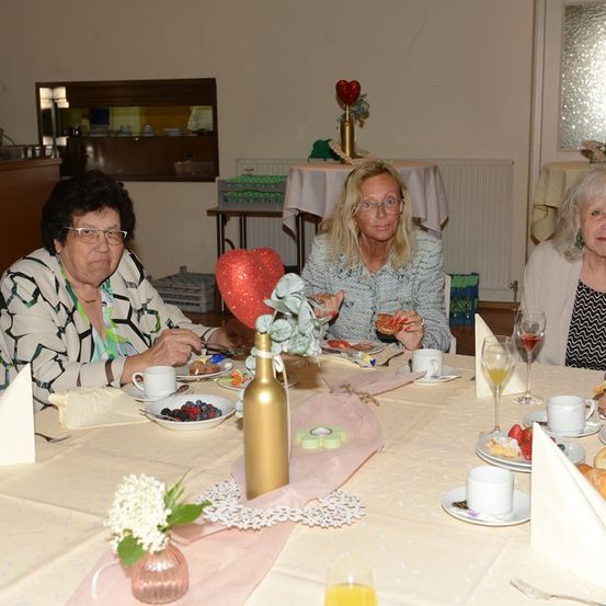 Drei Frauen sitzen an einem Tisch und genießen eine Mahlzeit. Der Tisch ist mit Tellern, Tassen und einer Flasche gedeckt. Eine Frau hält ein Stück Essen.