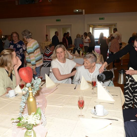 Eine Gruppe von Menschen versammelt sich in einem Raum, einige sitzen an Tischen, andere stehen und unterhalten sich. Eine Frau schenkt ein Getränk in ein Weinglas ein. Ein Tisch ist mit Blumen, Weingläsern und Tellern gedeckt.