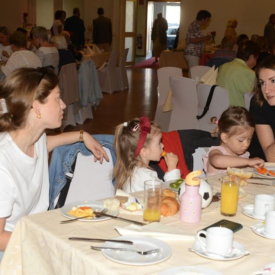 Eine Gruppe von Menschen, einschließlich Erwachsener und Kindern, versammelt sich in einem Essbereich. Zwei junge Mädchen essen an einem Tisch mit Tellern voller Speisen, Gläsern mit Orangensaft und anderen Gegenständen.