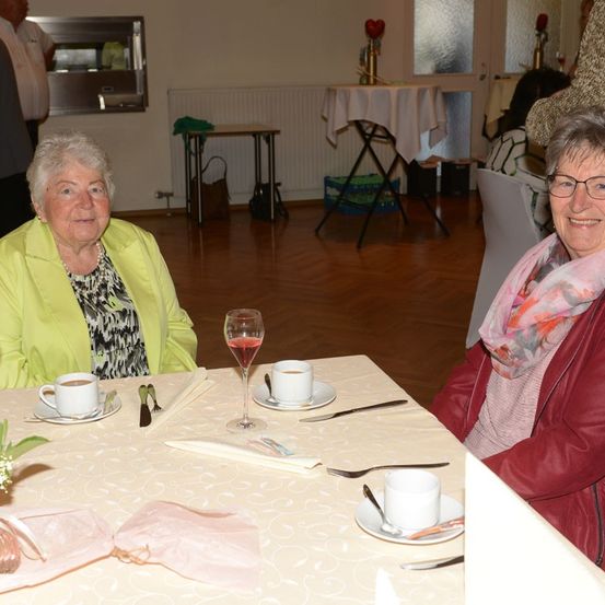 Zwei ältere Frauen sitzen an einem Tisch und lächeln, mit Tassen Kaffee und Wein vor ihnen, in einem Essbereich mit Holzböden und weißen Wänden.