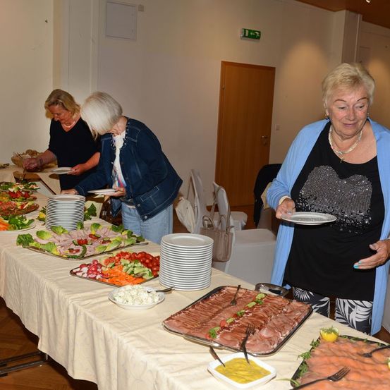Drei Frauen servieren Essen von einem Buffettisch mit verschiedenen Gerichten, einschließlich Fleisch, Gemüse und Salaten. Der Tisch ist mit einer weißen Tischdecke bedeckt.