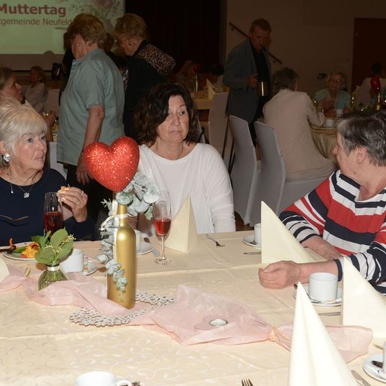 Eine Gruppe älterer Frauen und Männer sitzt an einem Tisch in einem Speisesaal, mit einem herzförmigen Mittelpunkt und Weingläsern. Der Tisch ist mit Tellern, Servietten und Besteck gedeckt. Eine Projektionsleinwand zeigt 'Muttertag' im Hintergrund.