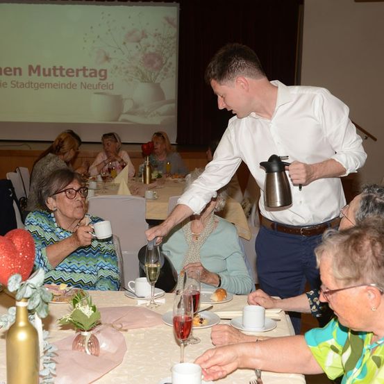 Ein Mann serviert Kaffee für ältere Frauen bei einem Muttertagsevent, mit einer Projektionsleinwand im Hintergrund, die den Titel der Veranstaltung anzeigt.