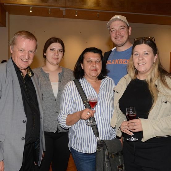 Fünf Personen lächeln und posieren für ein Foto in einer Innenumgebung. Die Frau in der Mitte hält ein Weinglas. Der Mann links trägt einen grauen Anzug und die Frau rechts eine beige Jacke.