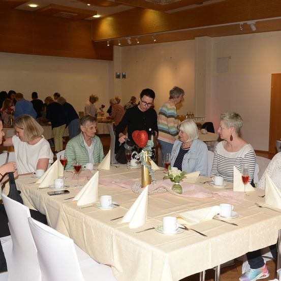 Eine Gruppe von Frauen sitzt an einem langen Tisch in einem Speisesaal, einige mit Weingläsern, andere mit Tassen und Untertassen. Eine Frau in einem schwarzen Shirt hält ein rotes Herz.