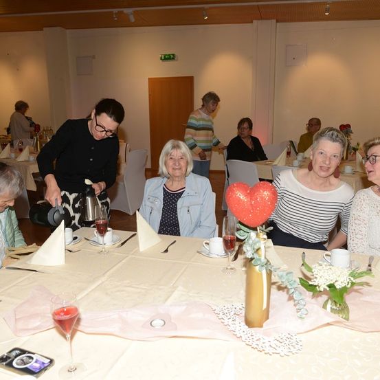 Eine Gruppe von Frauen sitzt an einem Tisch, eine Frau in Schwarz schenkt ein Getränk ein. Eine rote herzförmige Dekoration und Blumen sind auf dem Tisch.
