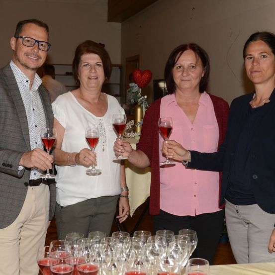 Vier Personen, zwei Männer und zwei Frauen, stehen zusammen und halten Gläser mit rotem Wein. Sie lächeln und scheinen ein Ereignis zu feiern. Hinter ihnen steht ein Tisch mit weiteren Weingläsern und einem dekorativen Herz.
