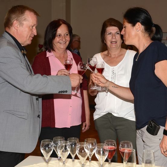 Vier Erwachsene, zwei Männer und zwei Frauen, stehen um einen Tisch mit Weinläsern, lächeln und halten Gläser mit rotem Wein.