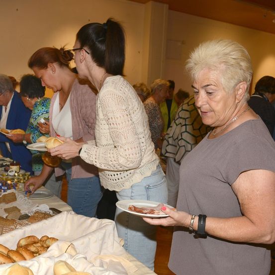 Mehrere Frauen stehen um einen Tisch mit Essen herum, darunter Brot, Gebäck und andere Gegenstände. Eine Frau hält einen Teller mit Essen und schaut eine andere Frau an.