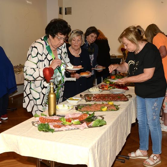 Mehrere Frauen stehen um einen Tisch mit verschiedenen Speisen, darunter Fleisch, Gemüse und Desserts. Eine Frau hält einen Teller mit Essen, und eine andere greift nach einer Schüssel. Der Tisch ist mit einer weißen Tischdecke bedeckt.