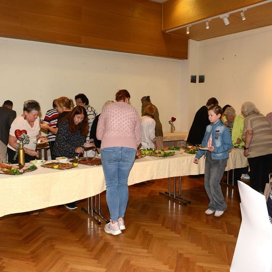 Eine Gruppe von Menschen versammelt sich um einen langen Tisch mit Essen, wahrscheinlich bei einem Buffet. Einige stehen, andere sitzen. Der Raum hat Holzböden und Wände.