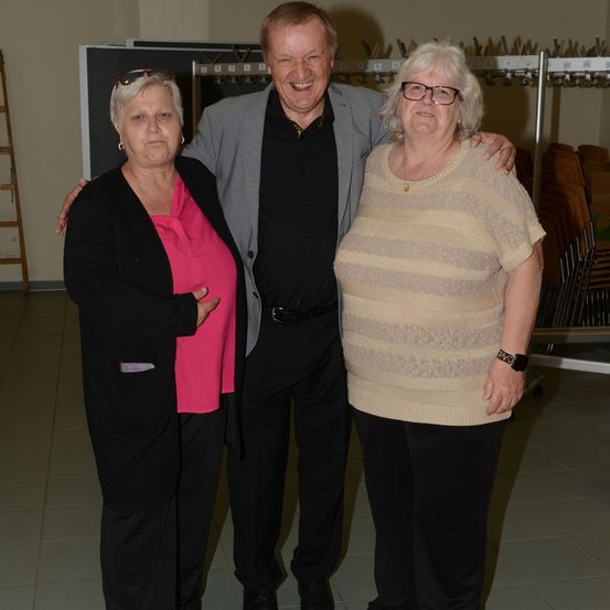 Drei Menschen stehen zusammen in einem Raum und lächeln für ein Foto. Der Mann hat seinen Arm um die beiden Frauen gelegt.