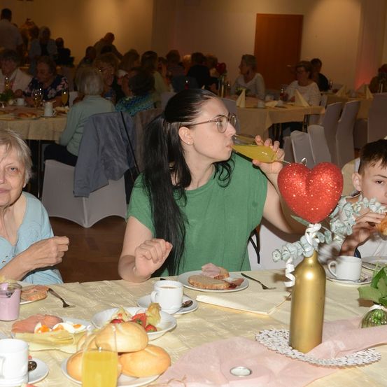 Eine Gruppe von Menschen, darunter eine ältere Frau und ein junges Mädchen, sitzen an einem Tisch und genießen eine Mahlzeit. Das junge Mädchen trinkt aus einem Glas, während die ältere Frau isst. Der Tisch ist mit Tellern, Tassen und einer Vase mit herzförmiger Dekoration gedeckt.