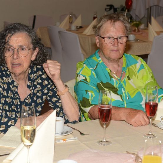 Zwei ältere Frauen sitzen an einem Tisch, eine mit Brille und Blumenhemd, die andere in einem grünen Blumenbluse. Sie haben Wein- und Kaffeegläser vor sich.