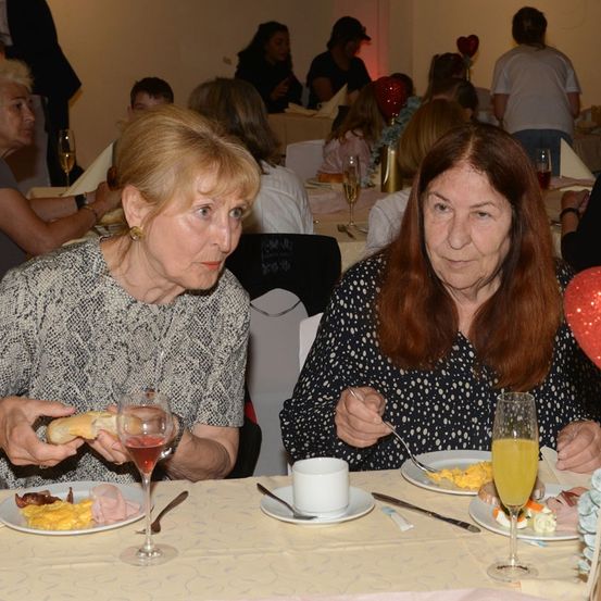 Zwei ältere Frauen sitzen an einem Tisch und frühstücken. Eine Frau hält ein Sandwich, während die andere Eier isst. Verschiedene Teller, Gläser und Bestecke stehen auf dem Tisch. Andere Personen sitzen an anderen Tischen im Hintergrund.