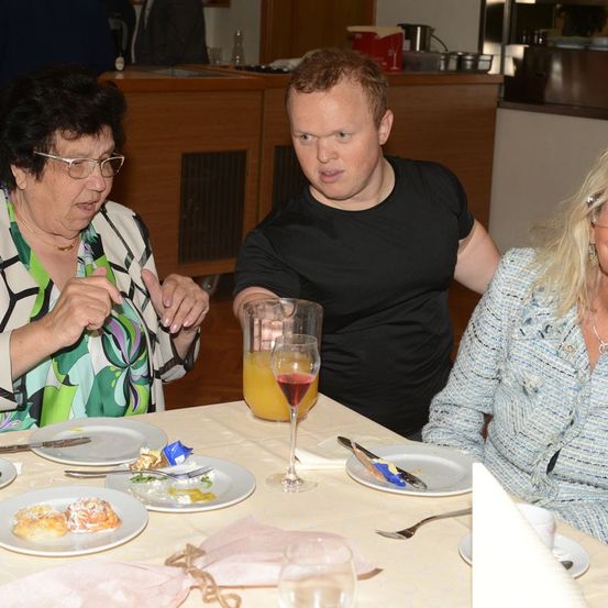 Drei Personen sitzen an einem Tisch, zwei Frauen und ein Mann, mit Essen und Getränken vor ihnen. Die Frau links trägt eine Brille und eine Halskette, der Mann trägt ein schwarzes T-Shirt und die Frau rechts trägt eine blaue Bluse.