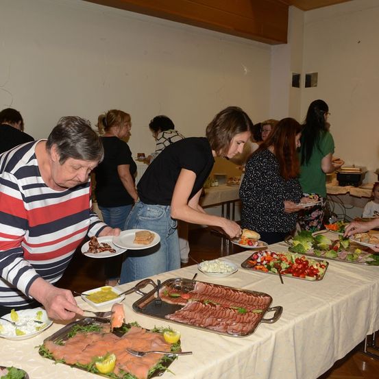 Eine Gruppe von Menschen versammelt sich um einen langen Tisch voller verschiedener Speisen, möglicherweise an einem Buffet. Einige nehmen Essen mit Tellern auf.