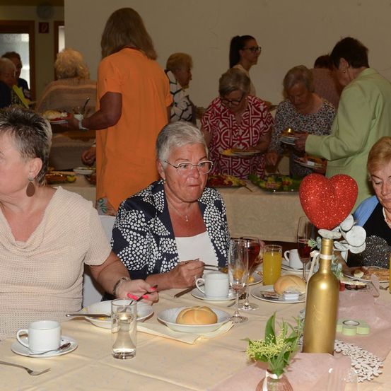 Eine Gruppe älterer Frauen versammelt sich um einen Tisch, wahrscheinlich bei einer sozialen Veranstaltung. Der Tisch ist mit Tellern, Gläsern und Brot gedeckt. Eine Frau trägt eine Brille und eine Halskette.