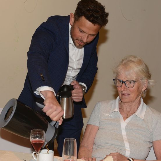 Ein Mann in einem blauen Anzug schenkt Rotwein in ein Glas für eine ältere Frau mit Brille und einem grauen Hemd.