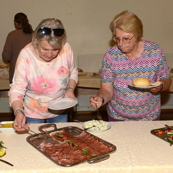 Zwei ältere Frauen servieren sich selbst Essen von einem Buffettisch, eine mit einem Teller Fleisch und die andere mit einem Sandwich.