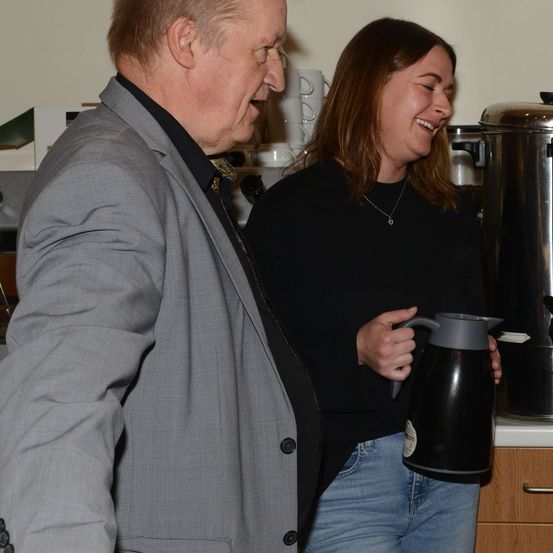 Ein Mann in einem grauen Anzug und eine Frau in einem schwarzen Shirt stehen in einer Küche, lächeln und halten eine Kaffeekanne.