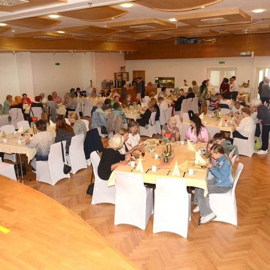 Ein großer Speisesaal mit vielen Menschen, die an Tischen sitzen, einige essen, während andere stehen und reden. Der Raum hat Holzböden und weiße Wände.
