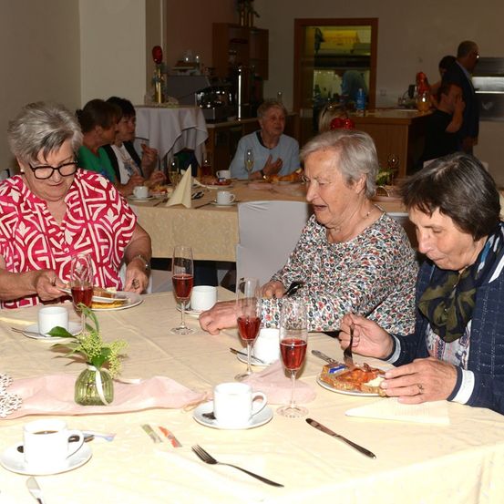 Eine Gruppe älterer Frauen sitzt an einem Tisch und isst und trinkt. Eine Frau trägt eine Brille und ein rotes Shirt, eine andere eine Bluse mit Blumenmuster. Weingläser, Teller und Servietten sind auf dem Tisch.
