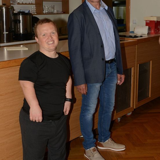 Zwei Männer stehen in einer Küche, einer in einem schwarzen T-Shirt und der andere in einem blauen Blazer und Jeans, lächeln für ein Foto.