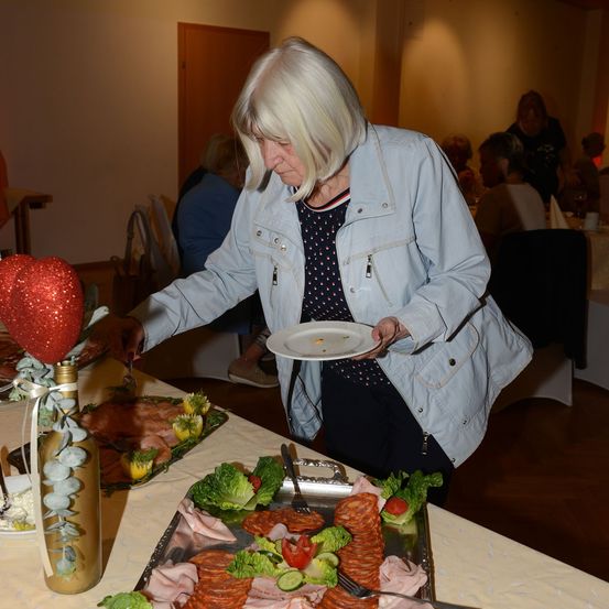 Eine ältere Frau serviert Essen von einem Buffettisch und hält einen Teller mit einem Stück Fleisch. Der Tisch ist mit verschiedenen Speisen gefüllt, darunter eine dekorative Flasche mit einem roten Herz obenauf.