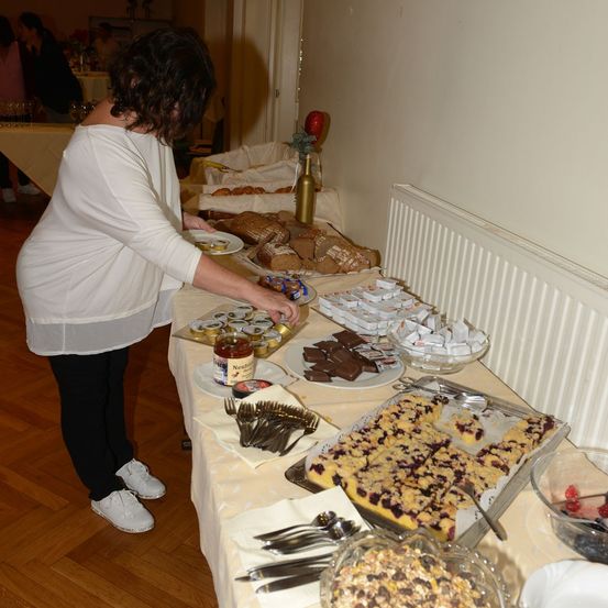 Eine Frau in einem weißen Shirt und schwarzen Hosen ordnet Essen auf einem Tisch an. Der Tisch ist mit verschiedenen Desserts gefüllt, darunter Brot, Schokoladen und Obst. Es gibt auch Schüsseln mit Beeren und eine Glasflasche mit Marmelade.