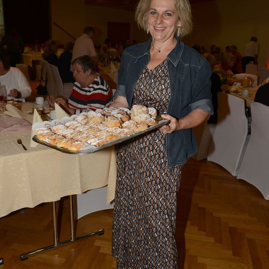 Eine Frau in einem gemusterten Kleid und einer Jeansjacke hält eine Tablett mit Gebäck bei einer gesellschaftlichen Veranstaltung.