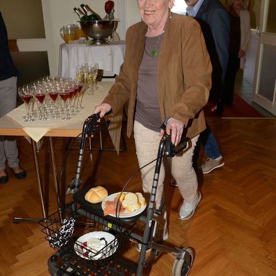Eine ältere Frau mit einem Rollator, lächelnd, geht an einem Tisch mit Weinläsern und Essen vorbei. Hinter ihr stehen ein Mann und eine Frau in der Nähe einer Tür.