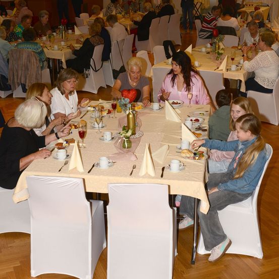 Eine Gruppe von Menschen, einschließlich älterer Frauen und Kindern, sitzt in einem Restaurant an einem Tisch und isst. Der Tisch ist mit einer Tischdecke bedeckt und hat Teller, Tassen und Weingläser. Der Raum hat Holzböden und weiße Stühle.