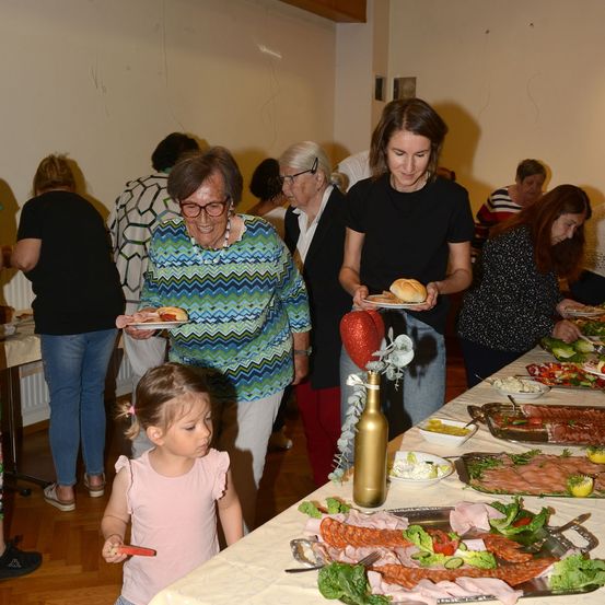 Eine Gruppe von Menschen, darunter Frauen und ein junges Mädchen, stehen um einen Tisch herum, der mit verschiedenen Speisen gefüllt ist, möglicherweise bei einem Buffet. Eine Frau hält einen Teller mit Essen und eine andere Frau hält einen Teller mit Brot. Ein junges Mädchen hält ein rotes Objekt.