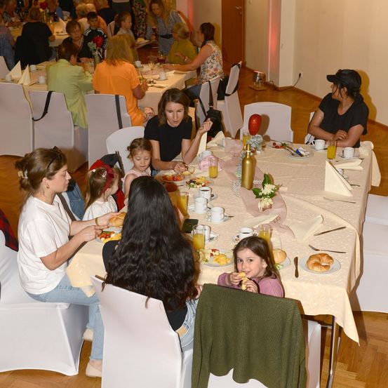 Eine Gruppe von Frauen und Kindern sitzt um einen langen Tisch, isst und trinkt. Der Tisch ist mit Tellern, Tassen und Servietten gedeckt. Der Raum hat Holzböden und weiße Wände.