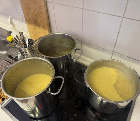 Drei Edelstahltöpfe mit gelblichem Inhalt stehen auf einem schwarzen Herd. Ein Topf hat einen Löffel darin. Hinter den Töpfen befindet sich eine weiße Fliesenwand und ein Holzbrett.