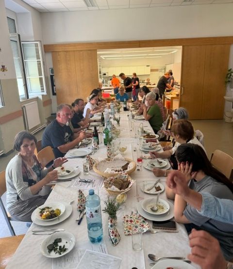 Eine Gruppe von Menschen sitzt um einen langen Tisch in einem Speisesaal, isst und trinkt. Der Tisch ist mit Tellern, Gläsern und verschiedenen Speisen gedeckt. Im Hintergrund stehen einige Personen, die möglicherweise Essen zubereiten. Der Raum hat Holzwände und eine weiße Decke mit Lichtern.
