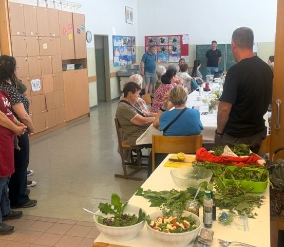 Eine Gruppe von Menschen ist um einen langen Tisch in einem Raum versammelt, möglicherweise bei der Essenszubereitung. Der Tisch ist mit verschiedenen Zutaten gefüllt, und einige Leute stehen, während andere sitzen. Es gibt Schränke und eine Uhr an der Wand.
