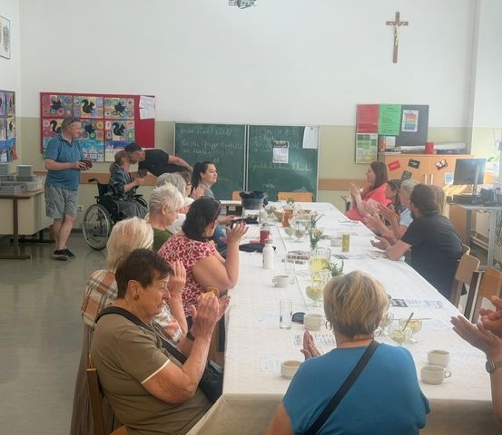Eine Gruppe älterer Menschen sitzt an einem langen Tisch in einem Raum, klatscht und lächelt. Ein Mann im Rollstuhl steht hinter dem Tisch. Ein Kreuz ist an der Wand.