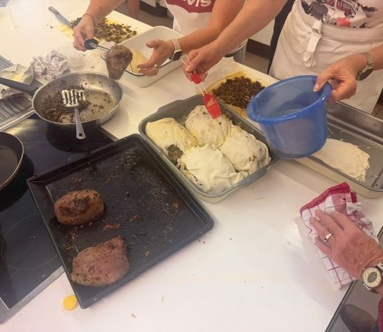 Mehrere Personen kochen in einer Küche, eine Person hält einen blauen Eimer und eine andere einen Schöpflöffel. Es gibt Tabletts mit Essen, eine Pfanne und ein Schneidebrett auf dem Tisch.