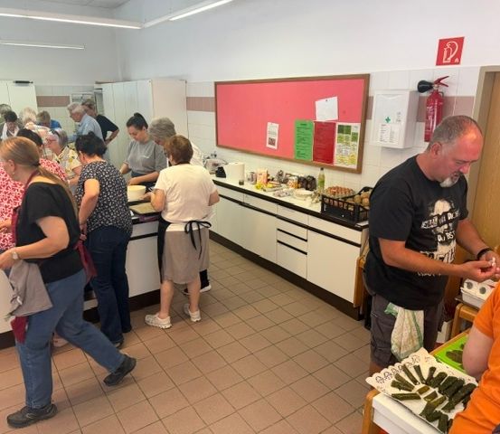 Eine Gruppe von Menschen befindet sich in einer Küche und bereitet Essen zu. Sie stehen um einen Tresen mit verschiedenen Zutaten und Utensilien. Ein Mann in einem schwarzen T-Shirt schaut sich etwas auf dem Tresen an.