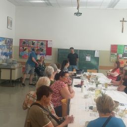 Eine Gruppe von Menschen, darunter eine ältere Frau im Rollstuhl, sitzt um einen langen Tisch in einem Raum. Ein Mann steht in der Nähe des Tisches und ein anderer Mann steht in der Nähe einer Tafel. Auf dem Tisch befinden sich verschiedene Gegenstände und ein Kreuz ist an der Wand montiert.